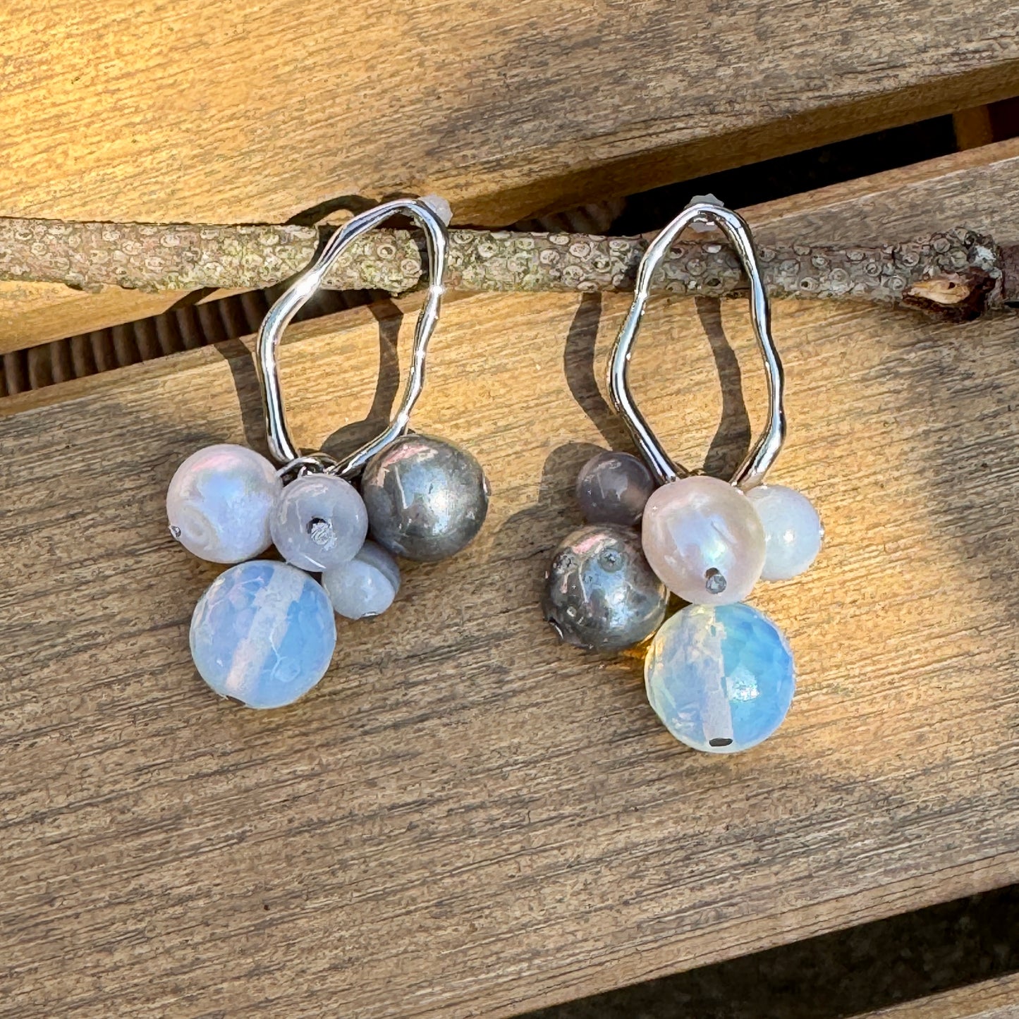 Baltic Bauble Earrings with hematite, agate, and freshwater pearl dangles on wavy rhodium hoops, styled on a light wood background