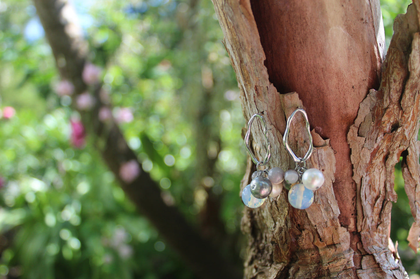 Baltic Bauble Earrings with hematite, opalite, and pearl beads on silver wavy hoops, hanging on a tree trunk with floral garden background