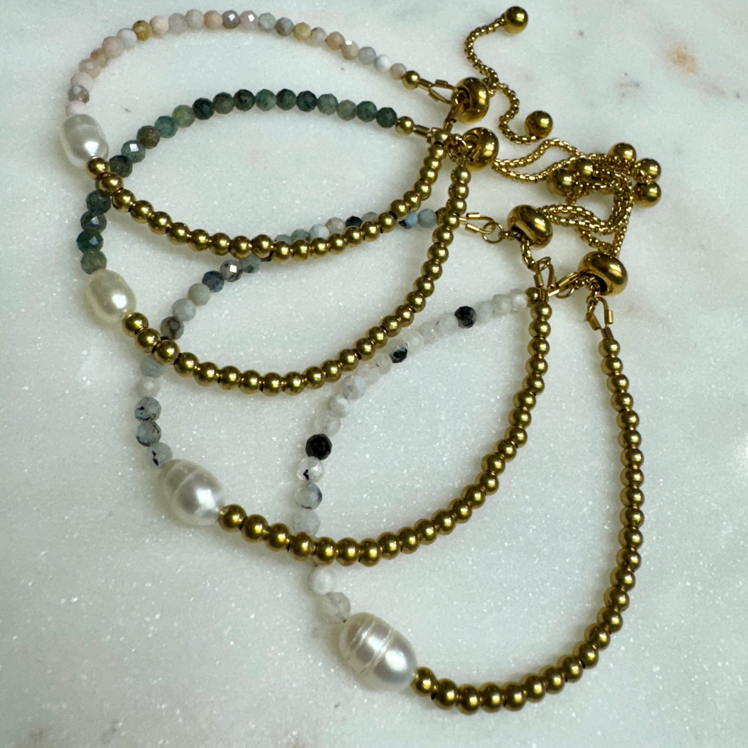 Flat lay of May & Moon’s adjustable beaded bracelets with gold accents, freshwater pearls, and faceted gemstone beads on marble — handmade, under $40, and perfect for layering or gifting.
