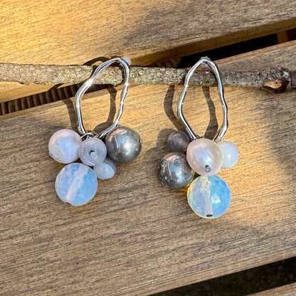 Baltic Bauble Earrings with hematite, agate, and freshwater pearl dangles on wavy rhodium hoops, styled on a light wood background
